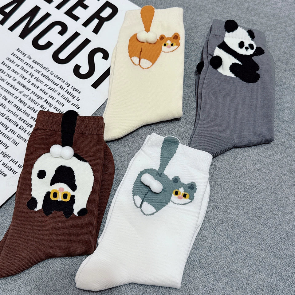 Four pairs of novelty socks arranged on a gray textured surface with a printed paper prop. The socks feature cute animal illustrations: a brown pair with a black-and-white cat, a cream pair with an orange cat, a gray pair with a panda, and a white pair with a gray rabbit and cat.