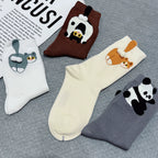 Four pairs of novelty socks arranged on a gray textured surface, each featuring a different cartoon animal face: a white rabbit, a brown cow, an orange fox, and a black-and-white panda. A printed paper is partially visible in the top left corner.
