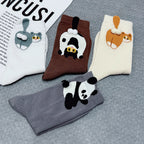 Four pairs of novelty socks arranged on a gray textured surface, each featuring a different cartoon animal applique: a blue rabbit, a brown panda, an orange cat, and a black-and-white panda. The socks come in white, brown, cream, and gray.